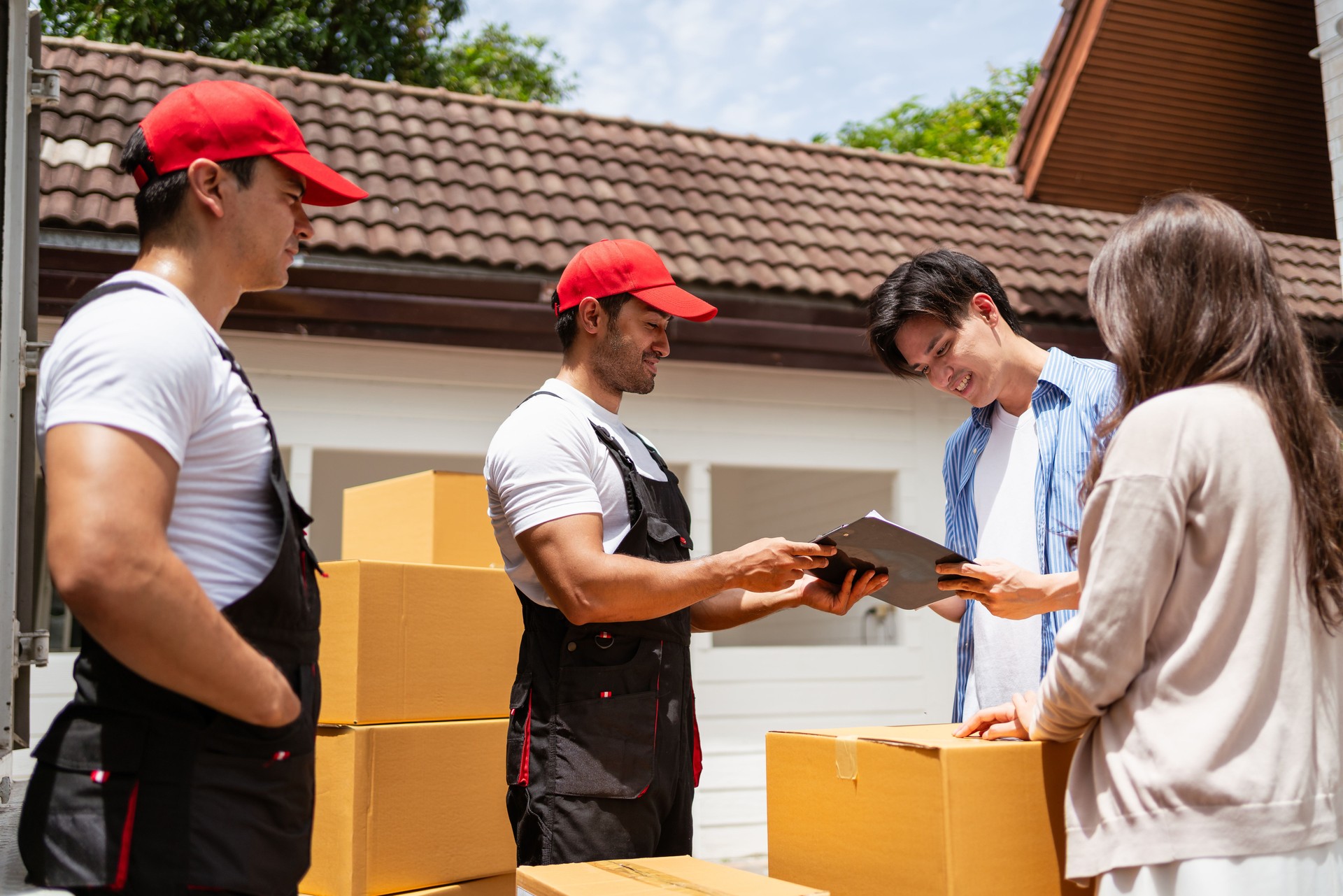 A couple signs the checklist of Asian and Caucasian workers in uniform unloading cardboard boxes from the truck. Delivery men unloading boxes to couple home. Professional delivery and moving service.
